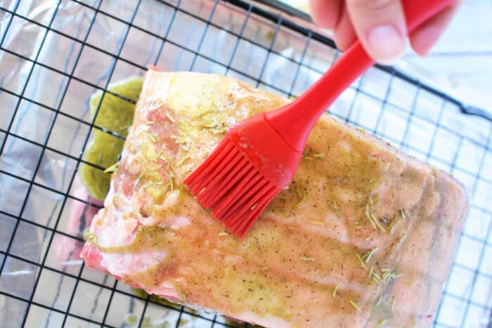 A close-up of a boneless rib roast on a metal cooling rack, covered in foil, being brushed with seasoning using a red silicone basting brush. The meat has a coat of a yellowish marinade with visible herbs and spices. A hand is holding the basting brush, perfect for any rib roast recipe.