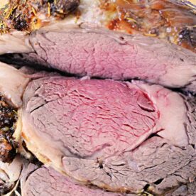 Close-up of a sliced medium-rare boneless rib roast revealing a juicy, pink interior surrounded by a well-cooked, brown outer layer. The meat is seasoned and garnished with some green herbs in the background.