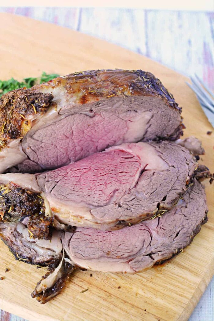 A beautifully sliced boneless rib roast on a wooden cutting board showcases medium-rare perfection in the center with a darker, well-done outer layer. Fresh herbs are sprinkled on top, accompanied by green garnish in the background. A fork is partially visible on the right side, completing this exquisite Boneless Rib Roast Recipe.