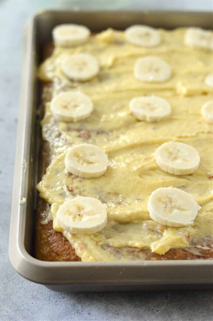 A rectangular cake in a baking pan is topped with creamy frosting and sliced bananas. Using a banana cake recipe with cake mix, the frosting is spread evenly across the surface, and the banana slices are artfully arranged on top. The pan sits on a light gray countertop.