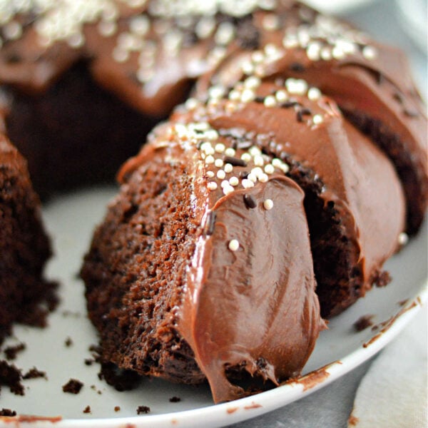 A chocolate bundt cake made with a cake mix with pudding, topped with rich chocolate frosting and white sprinkles, sits on a white plate. A slice has been cut out, revealing the moist interior enhanced by the pudding in the cake mix.