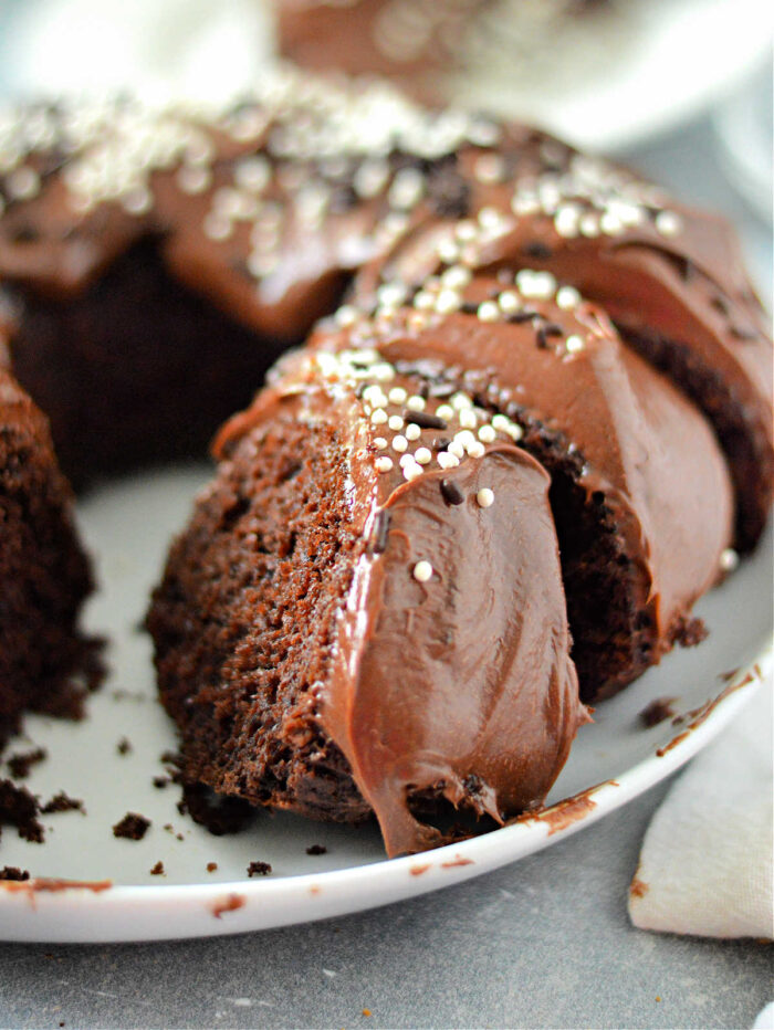 A chocolate bundt cake made with a cake mix with pudding, topped with rich chocolate frosting and white sprinkles, sits on a white plate. A slice has been cut out, revealing the moist interior enhanced by the pudding in the cake mix.