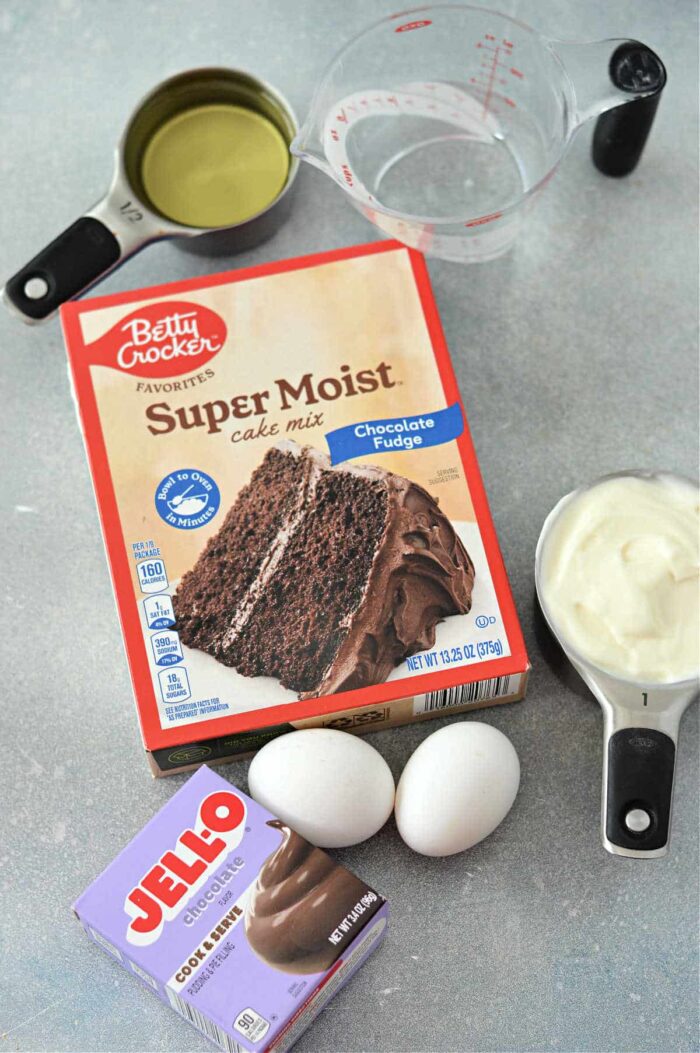 Image showing ingredients for a cake, featuring a box of Betty Crocker Super Moist chocolate fudge cake mix with pudding, a box of Jell-O chocolate pudding mix, two eggs, measuring cups with water and oil, and a bowl with a creamy white substance.