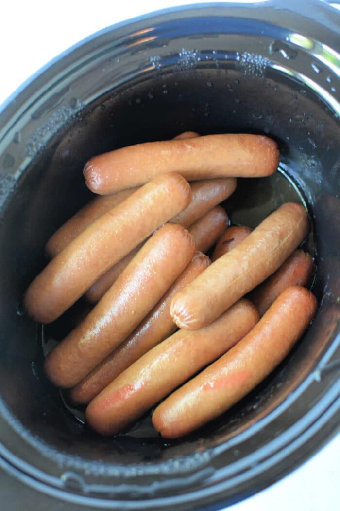 A black slow cooker filled with neatly arranged Slow Cooker Hot Dogs. The hot dogs are light brown and have a glossy appearance, indicating they have been recently cooked.