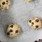 Four scoops of chocolate chip cookie dough are placed on a parchment-lined baking sheet, ready to be baked. The dough consists of light beige batter with visible chocolate chips. Perfect for an air fryer, this cookie dough sits in the center of the image, ready for a quick and easy treat.