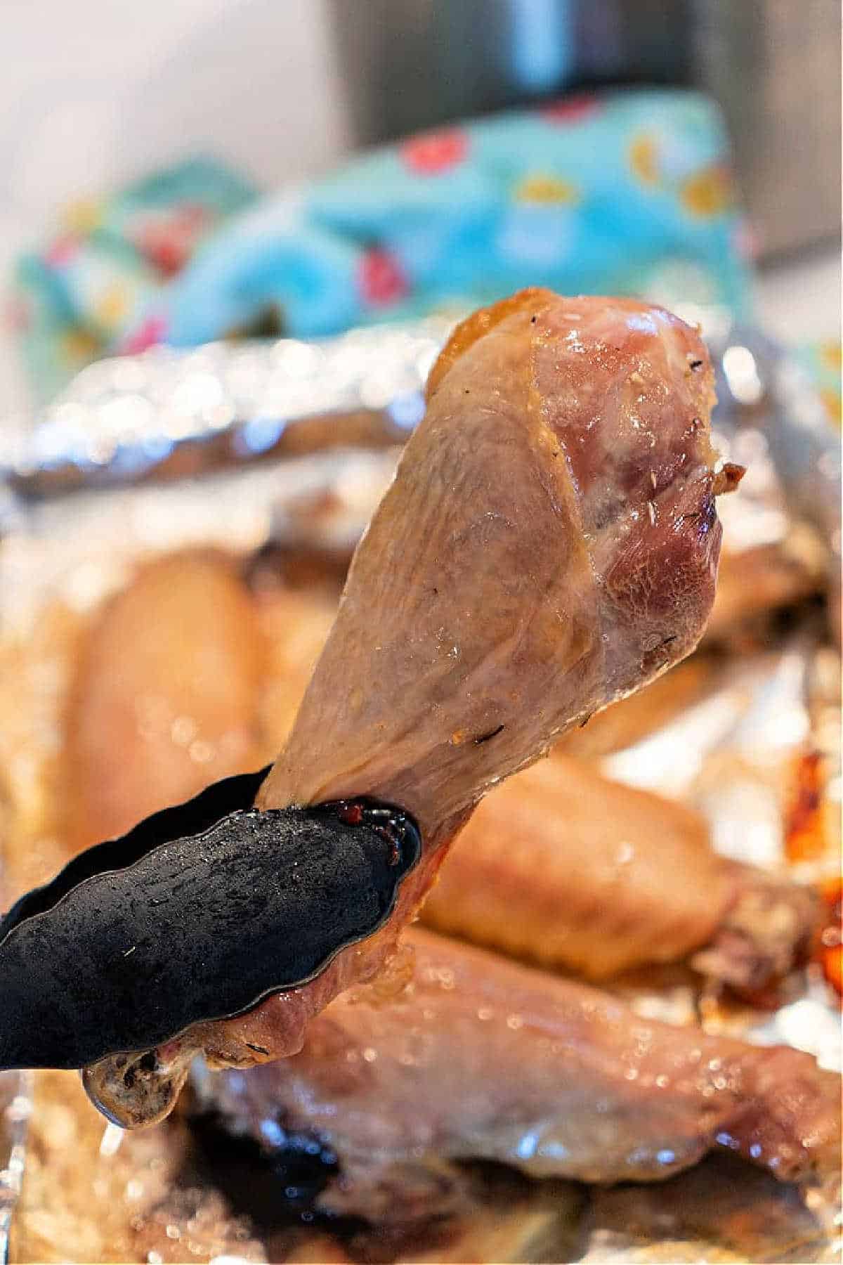 Tongs holding a cooked turkey drumstick above a tray with more baked turkey legs in the background.