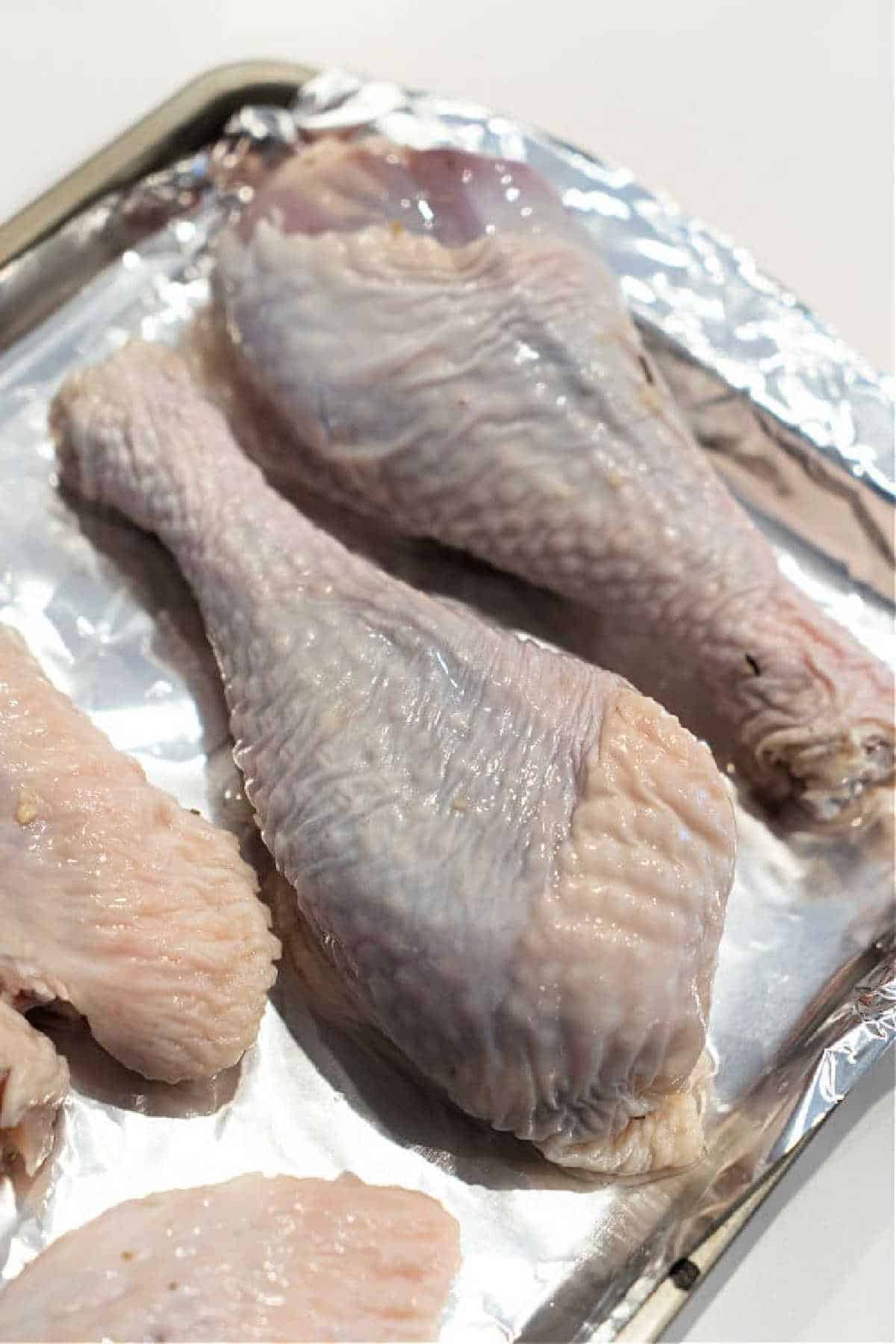 Raw chicken drumsticks and wings on a foil-lined baking tray, prepared similarly to baked turkey legs, ready for cooking.
