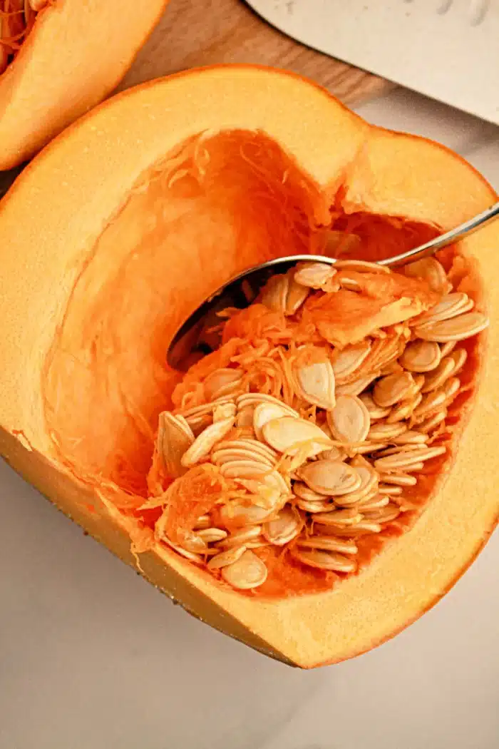 Close-up of a halved pumpkin with orange flesh, showing a spoon scooping out seeds from the center. The surface around the pumpkin appears to be a light-colored table. Picture preparing this for your next meal: pumpkin in slow cooker, complete with seeds and fibrous strands inside.