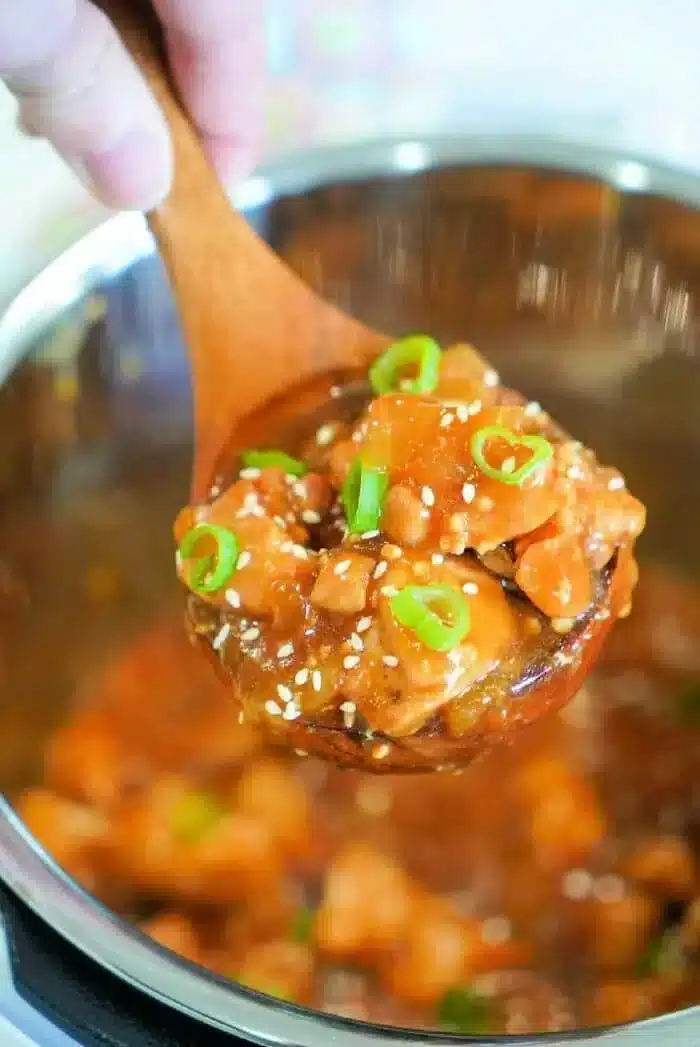 A close-up of a wooden spoon holding a serving of an Instant Pot Korean chicken dish with diced vegetables in a thick sauce. The dish is garnished with sesame seeds and sliced green onions. The background shows a pot containing the rest of the flavorful meal.