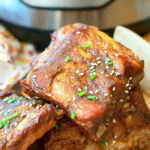 A stack of glazed pork ribs garnished with chives and sesame seeds sits temptingly in front of an Instant Pot, showcasing a mouthwatering instant pot teriyaki ribs recipe.
