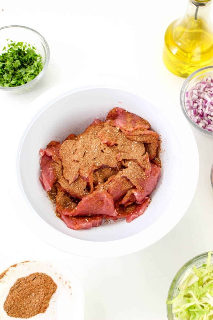 A white bowl contains raw chicken pieces seasoned with a brown spice mix, surrounded by bowls of chopped herbs, diced red onions, shredded lettuce, a small bowl of additional spices resembling the ones used in pork tenderloin tacos, and a bottle of olive oil on a white background.