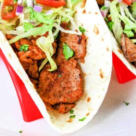 A close-up of a Pork Tenderloin Taco filled with seasoned meat, shredded lettuce, chopped tomatoes, diced red onions, and cilantro, all inside a soft flour tortilla. The taco is placed in a red taco holder. A lime wedge is also visible on top of the filling.