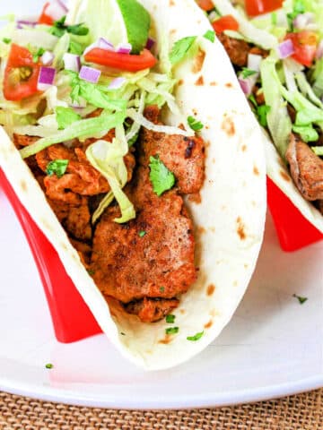 A close-up of a Pork Tenderloin Taco filled with seasoned meat, shredded lettuce, chopped tomatoes, diced red onions, and cilantro, all inside a soft flour tortilla. The taco is placed in a red taco holder. A lime wedge is also visible on top of the filling.