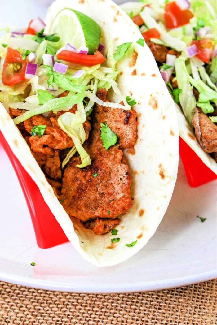 A close-up of a Pork Tenderloin Taco filled with seasoned meat, shredded lettuce, chopped tomatoes, diced red onions, and cilantro, all inside a soft flour tortilla. The taco is placed in a red taco holder. A lime wedge is also visible on top of the filling.