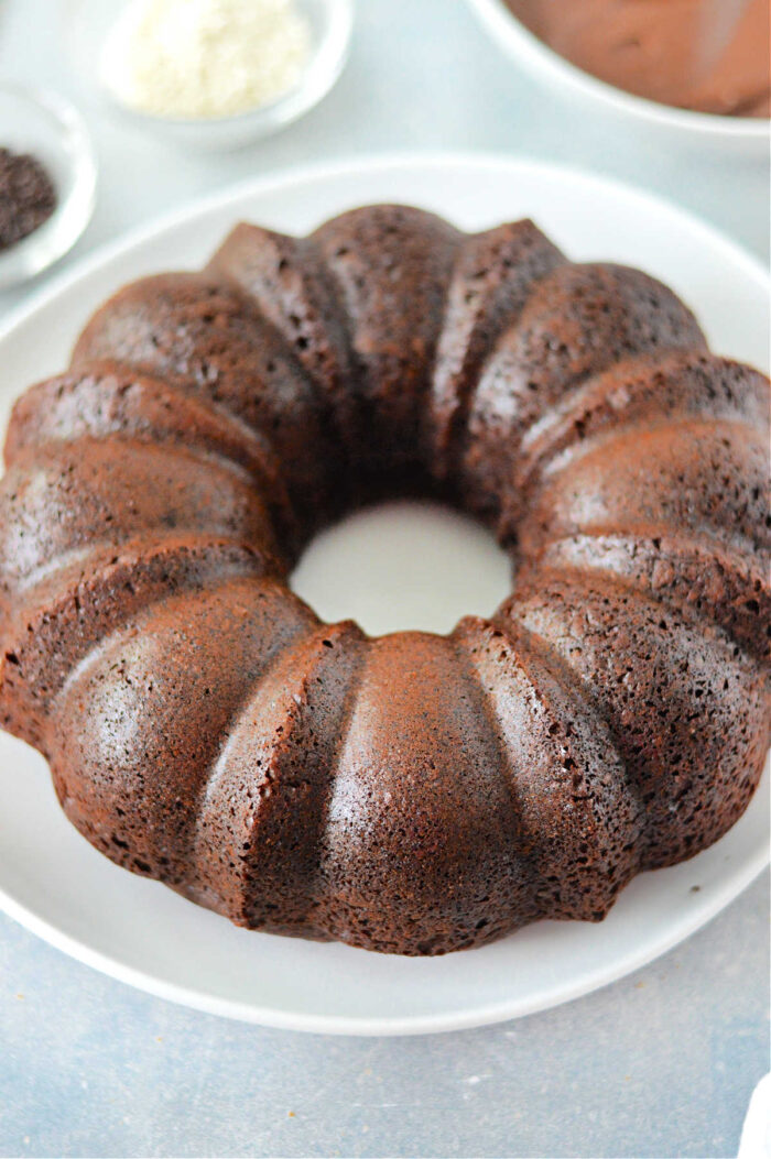 A chocolate bundt cake made with a cake mix with pudding is displayed on a white plate. The cake has a rich, dark brown color and a distinctive ridged shape characteristic of bundt cakes. The background includes blurred bowls containing various ingredients or toppings.