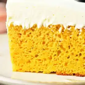 A close-up of a slice of pumpkin cake with yellow cake mix, featuring a thick layer of white frosting. The cake has a yellow, airy interior with visible bubbles and is placed on a beige plate.