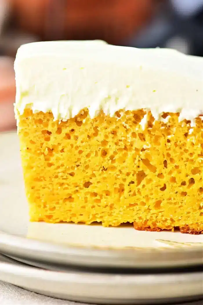 A close-up of a slice of pumpkin cake with yellow cake mix, featuring a thick layer of white frosting. The cake has a yellow, airy interior with visible bubbles and is placed on a beige plate.