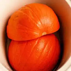 Two large, orange pumpkin halves placed inside a white slow cooker. The pumpkins appear fresh and slightly wet, fitting snugly within the cooker. The image suggests preparation for cooking or steaming the pumpkin in a slow cooker.