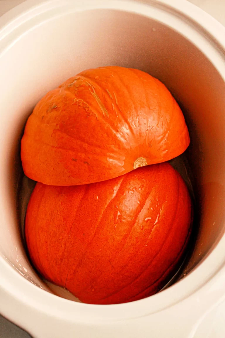 Two large, orange pumpkin halves placed inside a white slow cooker. The pumpkins appear fresh and slightly wet, fitting snugly within the cooker. The image suggests preparation for cooking or steaming the pumpkin in a slow cooker.