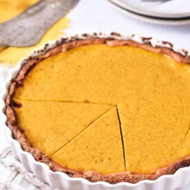 A sweet potato pie with a golden-brown crust and smooth filling sits in a white fluted pie dish. A slice has been cut and slightly separated from the rest of the pie. There are plates, forks, and a pie server in the background. A beige cloth is underneath the pie dish.