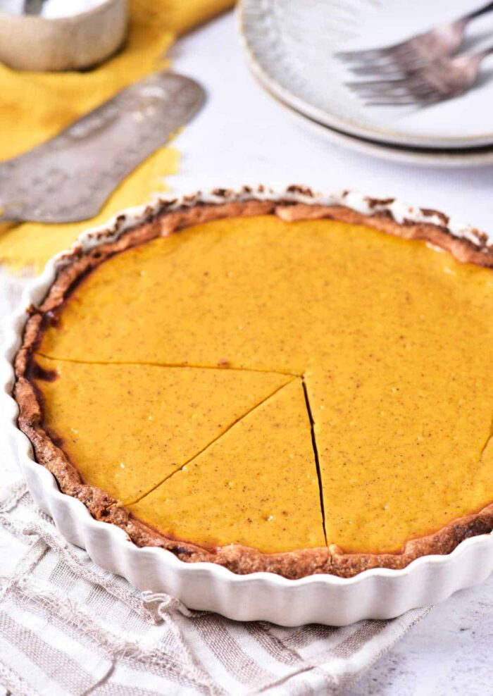 A pumpkin pie in a white pie dish is shown with a slice removed. The pie rests on a cloth napkin, with a silver serving utensil and some plates with forks in the background. The pie has a golden-brown crust and a smooth, orange filling, reminiscent of our Recipe for Sweet Potato Pie with Sweetened Condensed Milk.