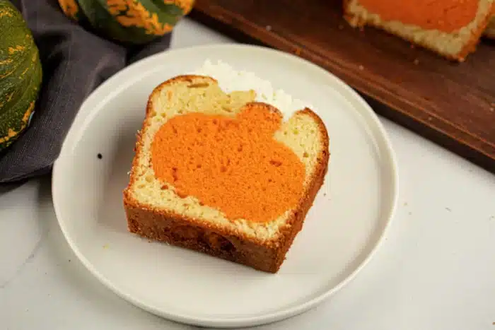 A slice of bread loaf on a white plate features an orange pumpkin shape inside the slice. In the background, a wooden cutting board holds another slice of the loaf, accompanied by a cloth and parts of green pumpkins, giving a hint that this might be a charming cake with an intriguing shape inside.