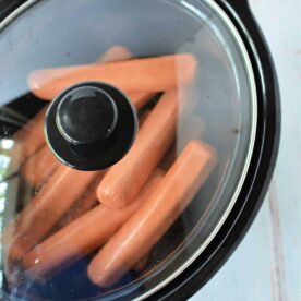 A black slow cooker with a glass lid contains several slow cooker hot dogs heating inside. The slow cooker is placed on a white surface with a faint pattern.