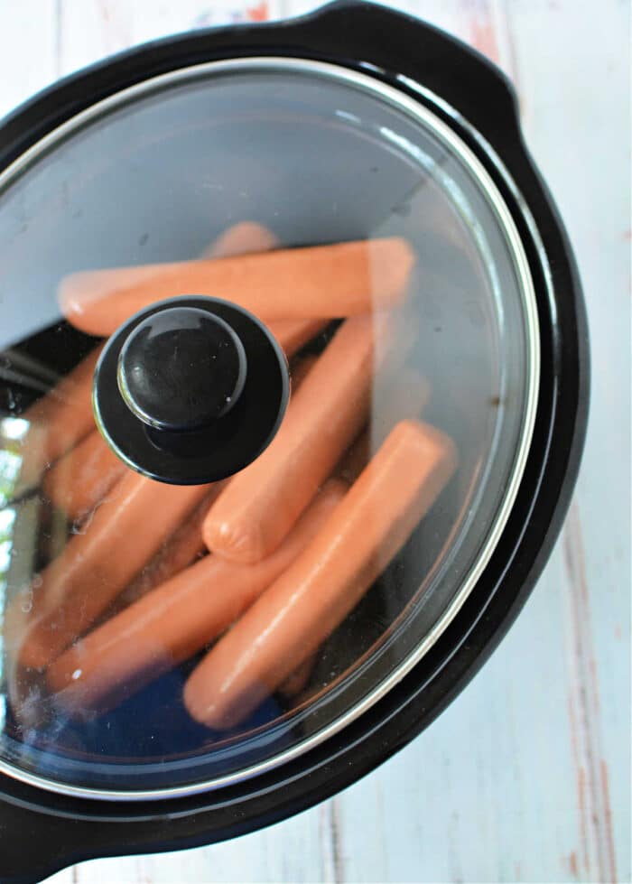 A black slow cooker with a glass lid contains several slow cooker hot dogs heating inside. The slow cooker is placed on a white surface with a faint pattern.