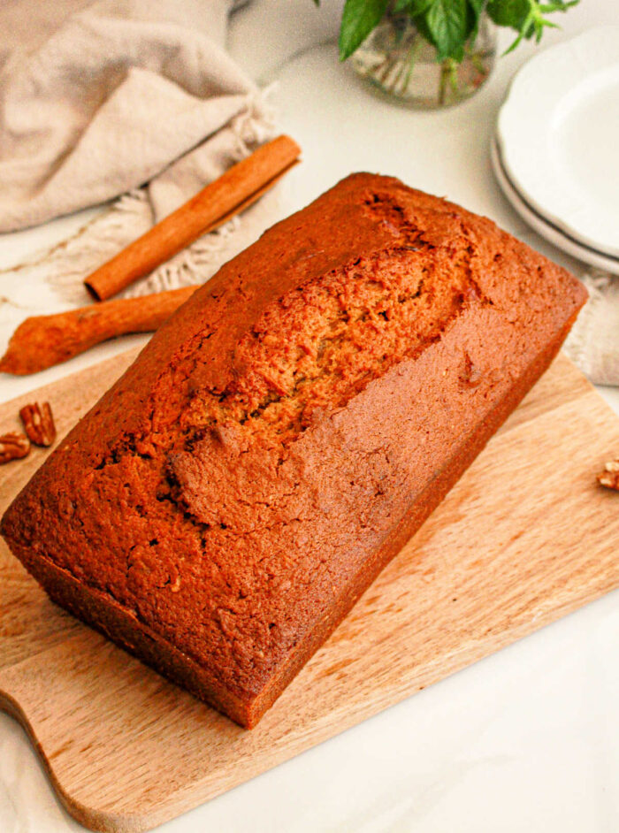 A loaf of freshly baked sweet potato bread with cake mix sits on a wooden cutting board. A cinnamon stick and some whole pecans are visible in the background, alongside a folded beige cloth and a stack of white plates.