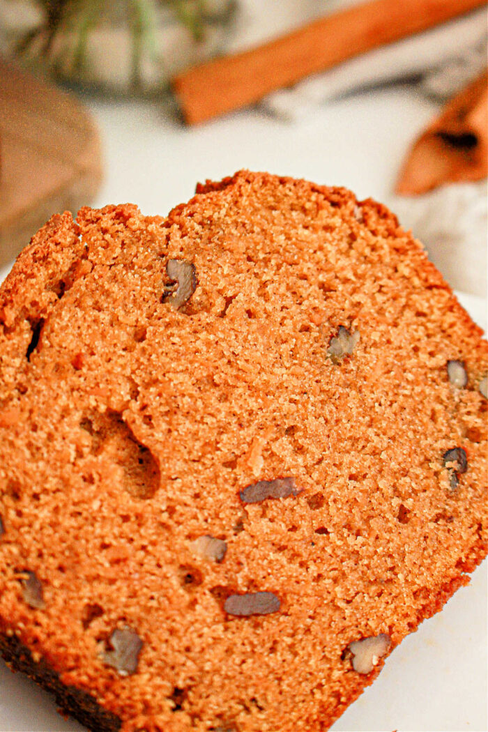 A close-up shot of a slice of pumpkin bread. The bread has a moist texture with visible pieces of nuts embedded throughout, reminiscent of sweet potato bread with cake mix. The background includes blurred elements hinting at additional ingredients or kitchen utensils.