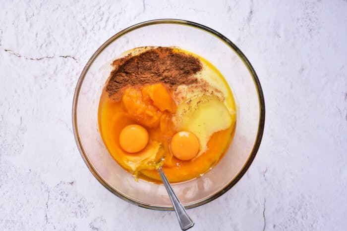 A glass bowl containing two raw eggs, mashed sweet potatoes, condensed milk, and spices like cinnamon and nutmeg sits on a white marbled surface. A spoon is also placed inside the bowl.