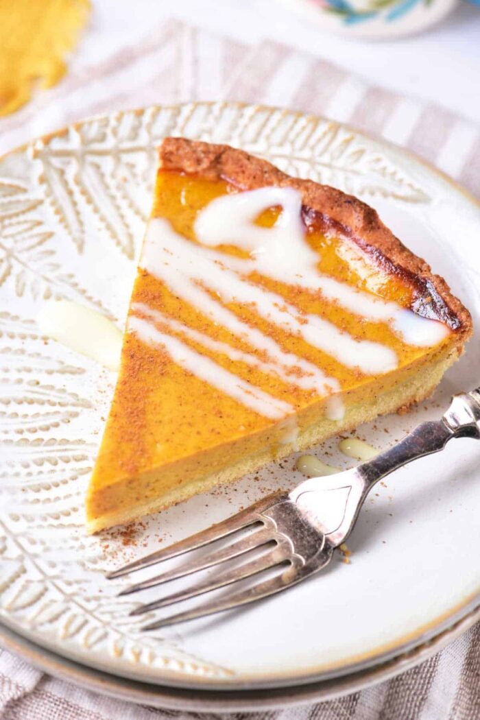 A slice of pumpkin pie topped with a drizzle of white icing on a decorative white plate. A silver fork rests beside the pie slice on the plate, evoking thoughts of a comforting recipe for sweet potato pie with sweetened condensed milk. The plate sits on a cloth with a woven pattern.