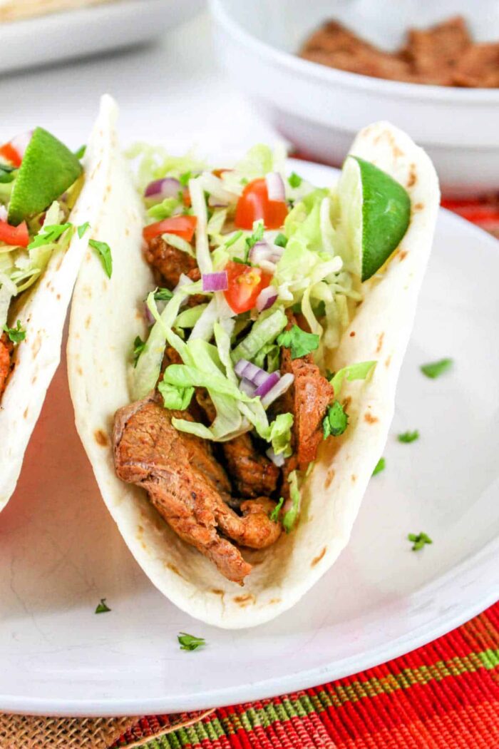 A close-up of a pork tenderloin taco filled with grilled pieces of meat, shredded lettuce, chopped tomatoes, and diced purple onions, garnished with a lime wedge. The taco is placed on a white plate with part of another taco visible in the background.