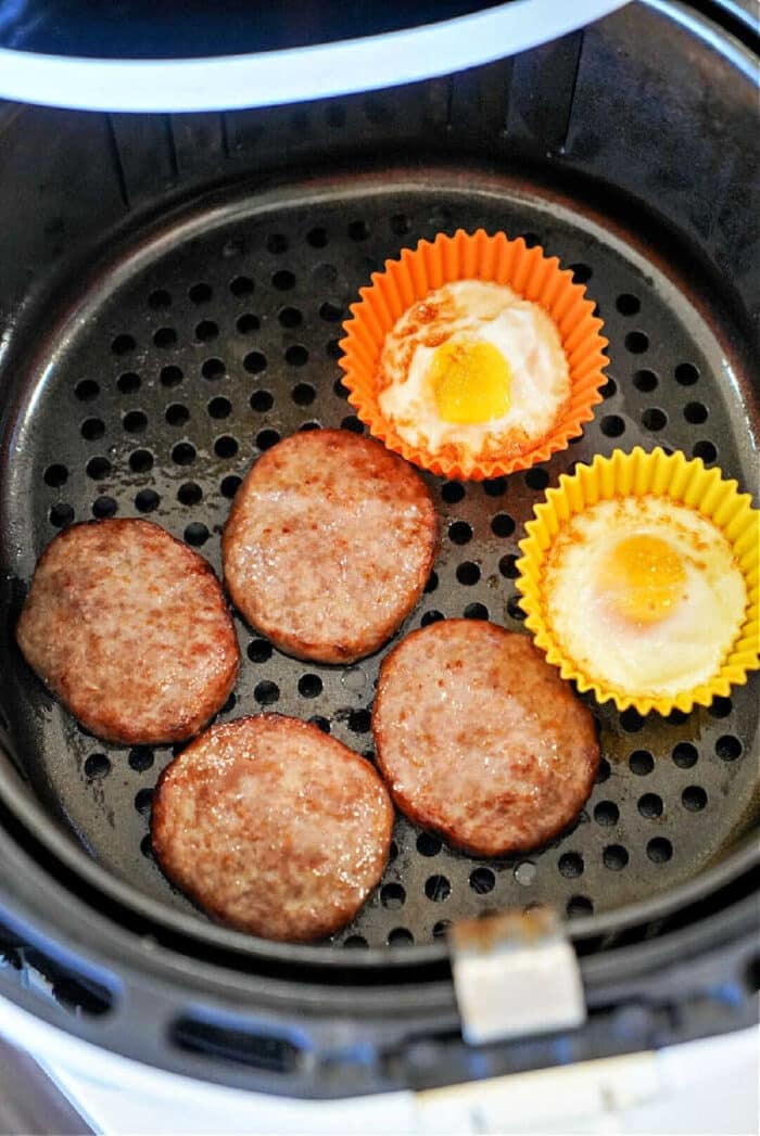 Five cooked sausage patties in air fryer and two eggs in silicone cups rest inside the basket, ready to serve.