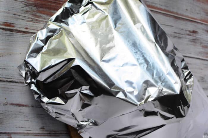 A piece of food, possibly involving a boneless rib roast recipe, is wrapped in crinkled aluminum foil on a wooden surface. The aluminum foil reflects light and covers the object completely. The wood surface has a whitewashed finish with visible grain patterns.