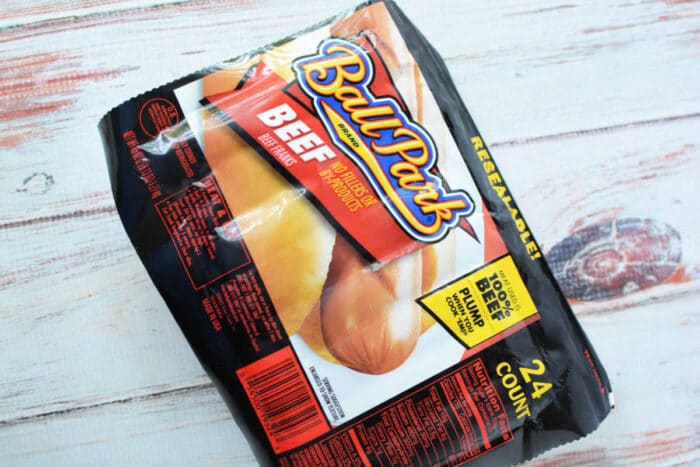 A package of Ball Park brand beef hot dog buns sits on a wooden surface. The packaging shows hot dogs and reads "BEEF - Made with 100% Beef Hot Dogs." Perfect for making slow cooker hot dogs, the package contains 24 individually wrapped hot dogs.