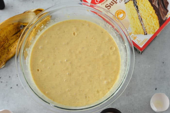 A glass bowl filled with cake batter sits on a gray surface. Next to the bowl, there is a banana peel, a box featuring an enticing banana cake recipe with cake mix, and egg shells. The cake mix box shows a yellow cake with chocolate frosting.