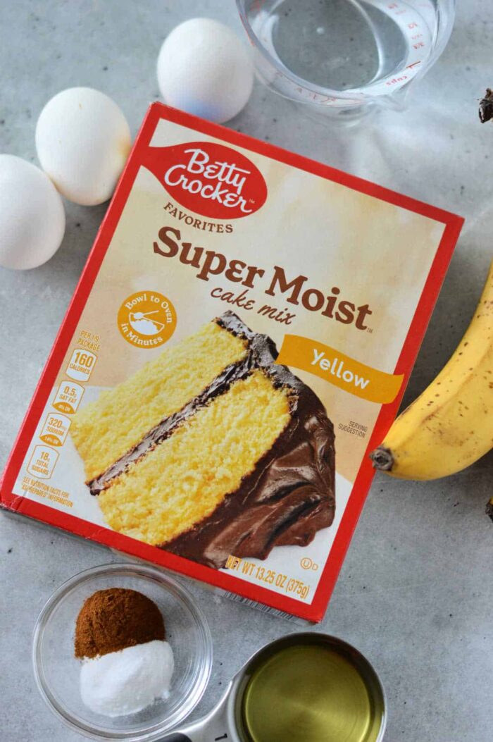 A box of Betty Crocker Super Moist yellow cake mix is on a countertop. Around it are three eggs, a peeled banana, a cup of water, a small cup of oil, a bowl of cinnamon, and a bowl of baking soda—everything you need for an effortless banana cake recipe with cake mix.