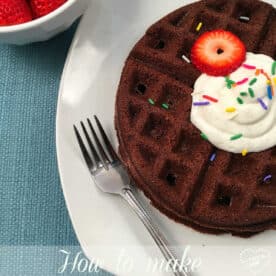 Brownie waffles topped with whipped cream, sprinkles, and a strawberry slice on a plate, served with a fork and a bowl of fresh strawberries.
