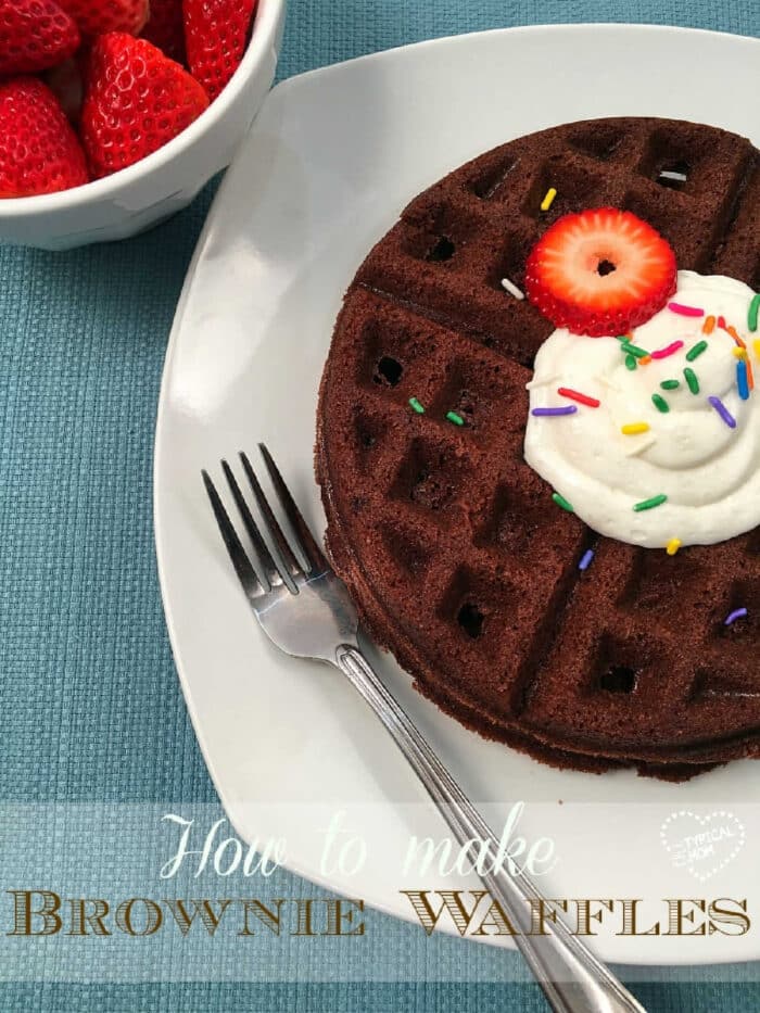 Brownie waffles topped with whipped cream, sprinkles, and a strawberry slice on a plate, served with a fork and a bowl of fresh strawberries.