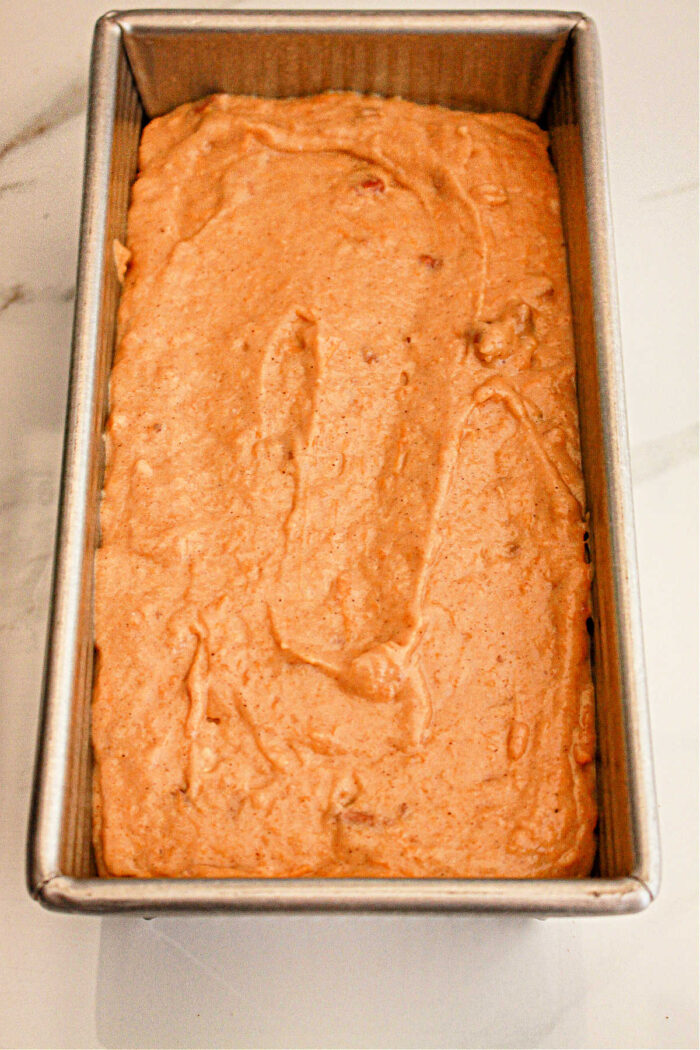 A metal loaf pan filled with unbaked sweet potato bread batter made with cake mix sits on a white surface. The batter appears smooth and evenly spread within the pan, ready for baking.