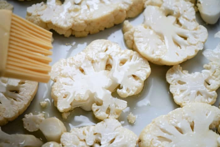 A brush spreads oil on sliced cauliflower pieces arranged on a baking sheet, preparing them for delicious cauliflower steak air fryer cooking.