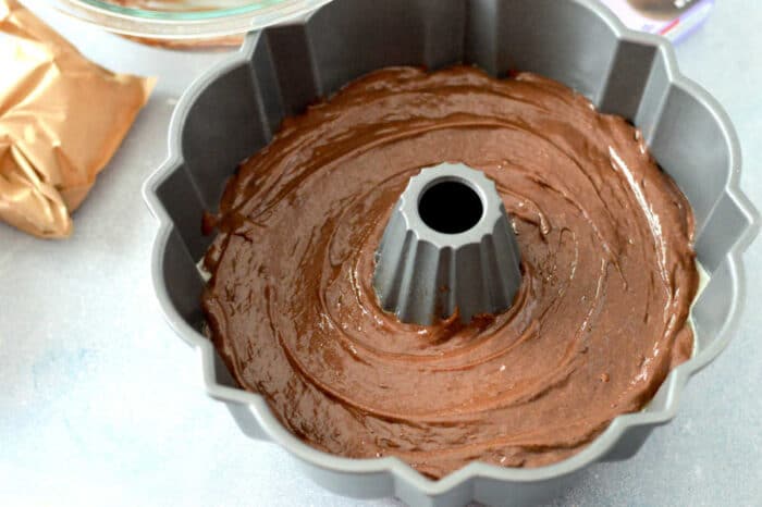 A bundt pan filled with chocolate cake mix with pudding is placed on a gray countertop. Surrounding the pan are various baking supplies, including a glass mixing bowl and a brown paper bag.