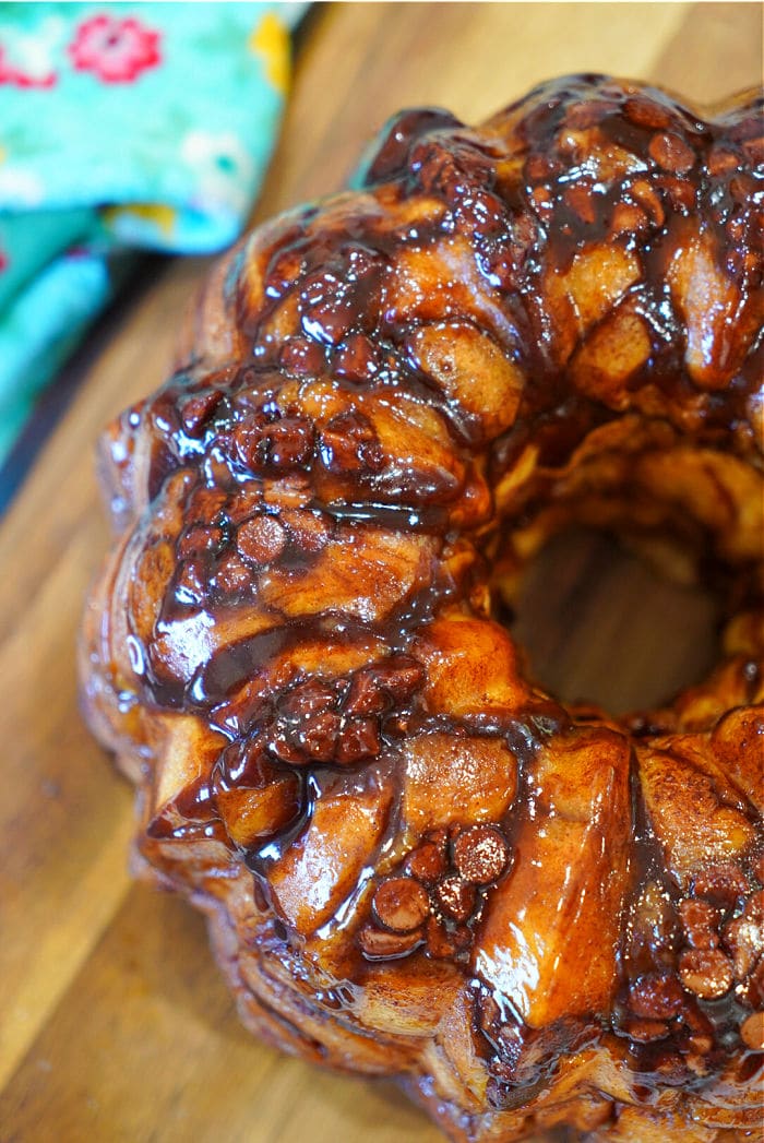 Close-up of glazed rhodes monkey bread with cinnamon and chocolate on a wooden surface, colorful cloth in background.
