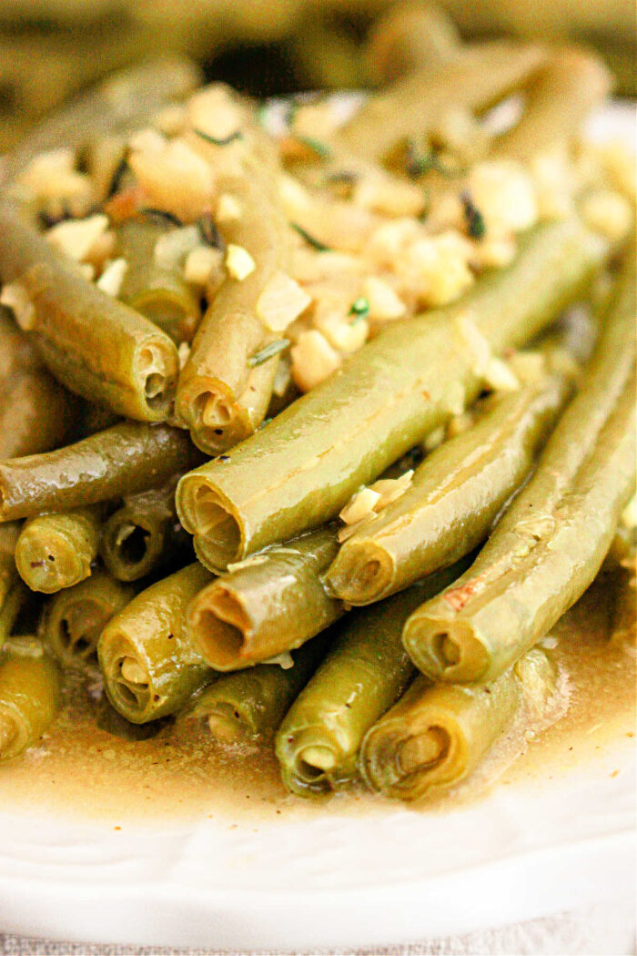 A close-up of crockpot-cooked green beans topped with finely chopped garlic and herbs. The green beans are slightly glossy from the seasoning and arranged in a pile on a white plate.