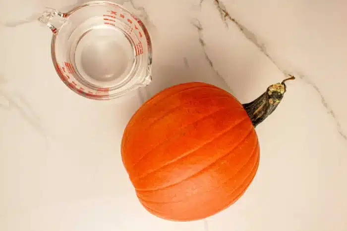 A small, round orange pumpkin with a stem lies next to a clear measuring cup filled partially with water on a white, marble-like surface, perfect for preparing pumpkin in a slow cooker.