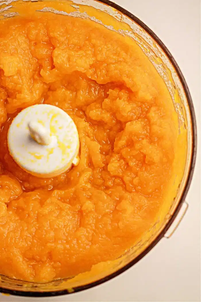 A food processor bowl filled with smooth, bright orange puree from pumpkin cooked in a slow cooker. The puree is evenly blended, and the white blade of the food processor is visible in the center. The sides of the bowl are coated with some of the mixture.