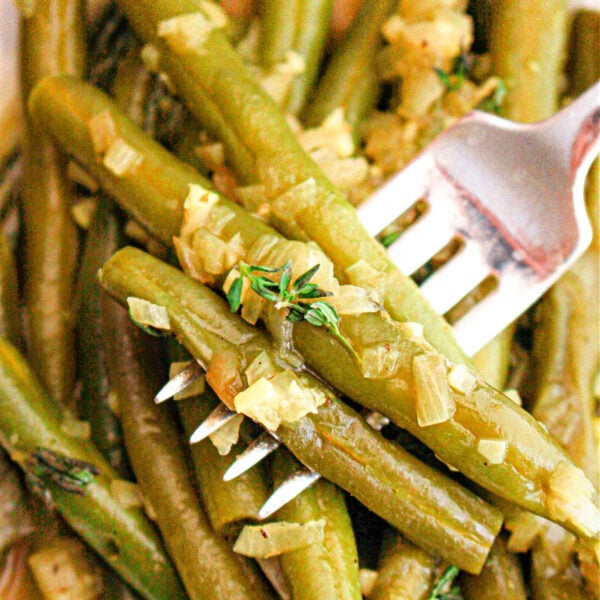 A close-up image of crockpot-cooked green beans seasoned with chopped onions and herbs. A fork is piercing several of the green beans, and the dish appears to be served on a white plate.