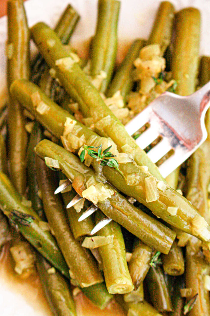 A close-up image of crockpot-cooked green beans seasoned with chopped onions and herbs. A fork is piercing several of the green beans, and the dish appears to be served on a white plate.