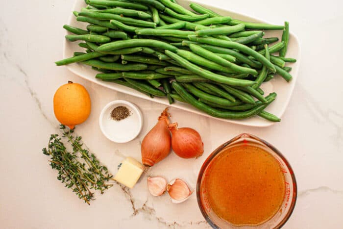 A display of ingredients on a white marble surface: a rectangular dish filled with fresh green beans, perfect for the crockpot, a whole lemon, small glass bowls with black pepper and broth, two shallots, a pat of butter, two garlic cloves, and a few sprigs of thyme.